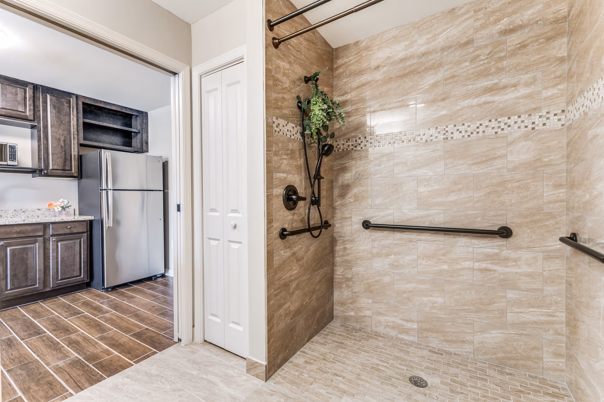 A spacious walk-in shower with beige tiled walls and floor, featuring a horizontal decorative tile strip and black grab bars. The shower has a black handheld showerhead with a small green plant hanging from it. Adjacent to the shower is a doorway leading to a kitchen area with dark wooden cabinets, a stainless steel refrigerator, and a microwave on the countertop.