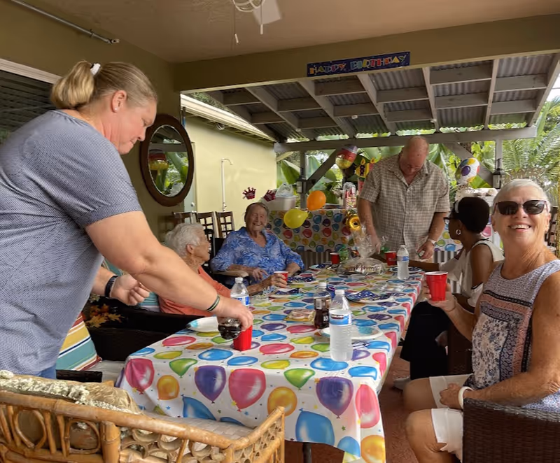 A group of elderly people and a caregiver gathered around a table decorated with a colorful balloon-themed tablecloth for a birthday celebration. The setting is a covered outdoor patio with birthday decorations including balloons and a 'Happy Birthday' banner. One woman is pouring a drink into a red cup while others are seated, smiling and enjoying the event.