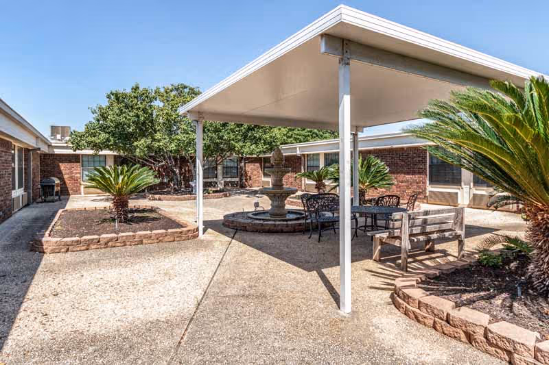 Outdoor courtyard area with a covered seating space including a table and chairs, a wooden bench, a central water fountain, and landscaped garden beds with palm plants and trees, surrounded by single-story brick buildings under a clear blue sky.