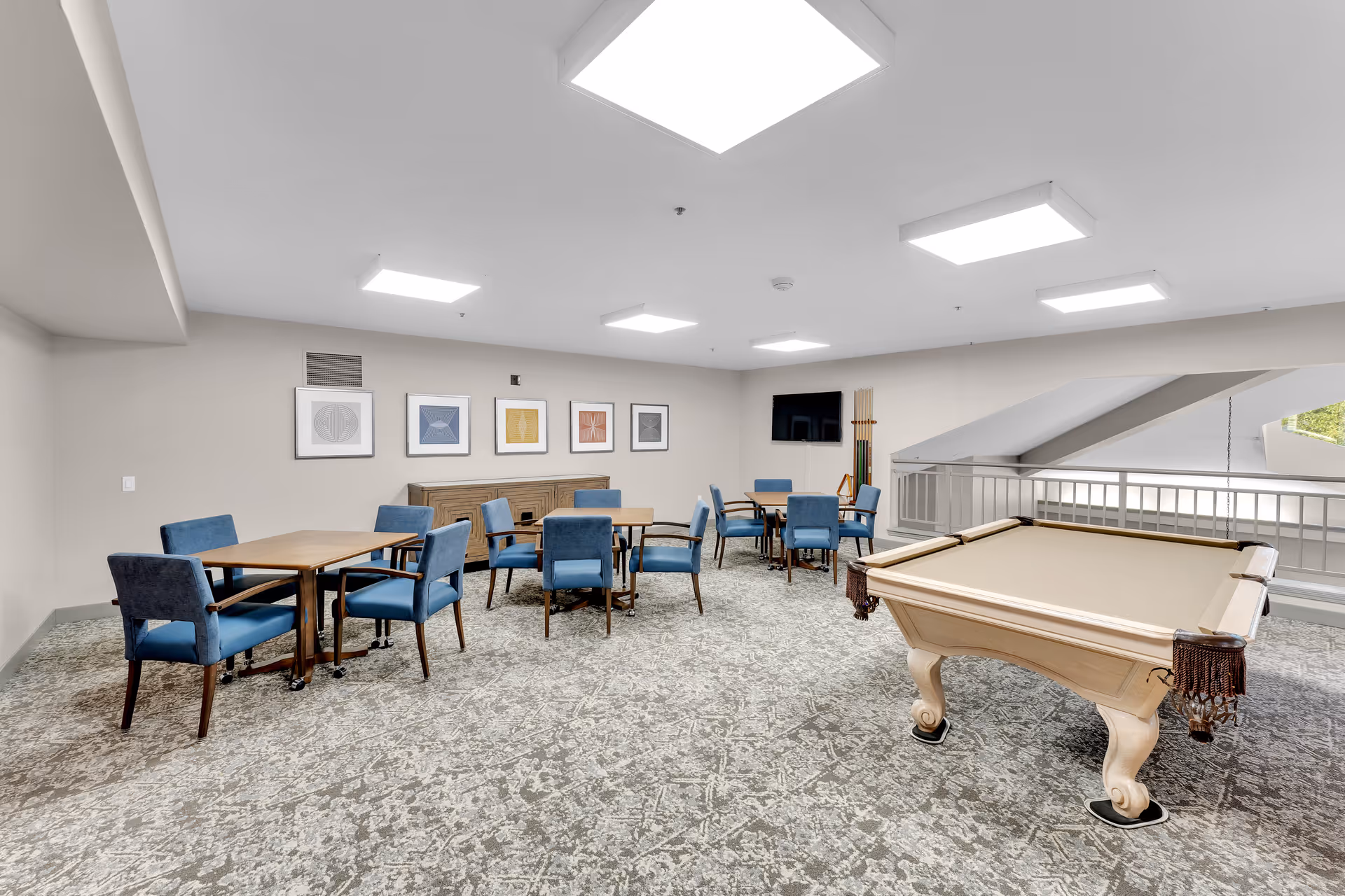 A well-lit recreational room with a beige pool table on the right and several wooden tables with blue upholstered chairs on the left. The room has a patterned carpet, white walls adorned with framed artwork, a flat-screen TV mounted on the far wall, and a rack holding pool cues. A railing and large windows are visible in the background.