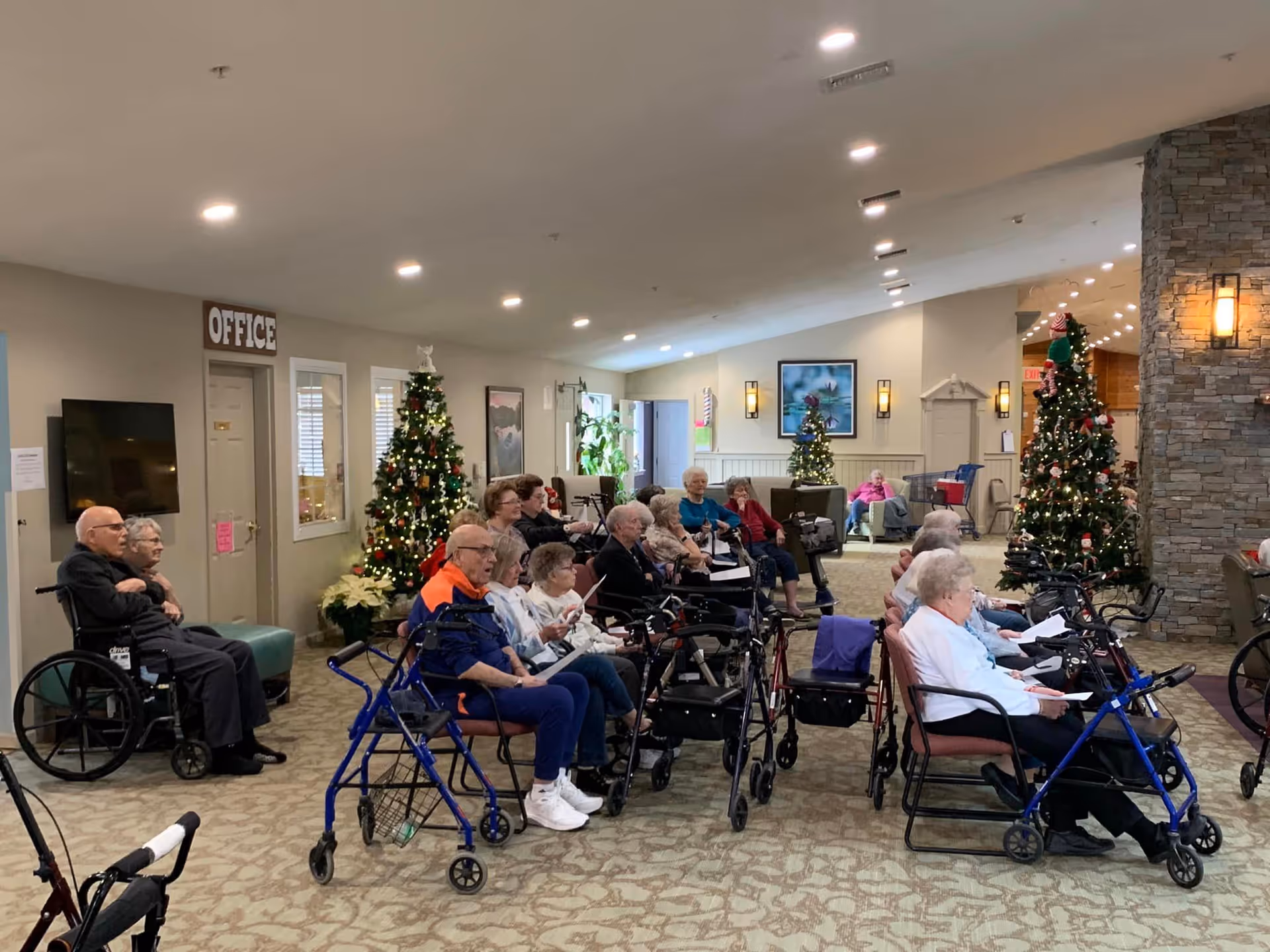 A group of elderly people seated in chairs and wheelchairs in a spacious, well-lit common area decorated with Christmas trees and holiday decorations. The room has a stone pillar with a wall sconce light, and a sign labeled 'OFFICE' is visible on the left side. The atmosphere appears calm and communal.