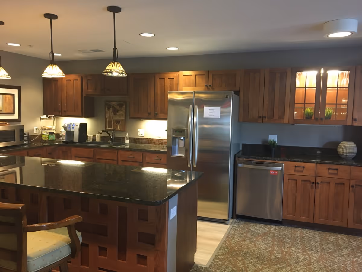 A modern kitchen with wooden cabinets, a large black granite island countertop with a chair, stainless steel refrigerator and dishwasher, pendant lights hanging from the ceiling, and decorative plants and artwork.