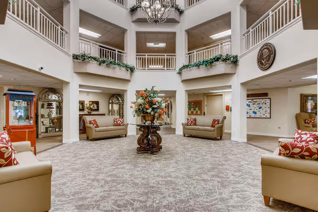 Spacious and well-lit common area in a senior living facility with beige sofas adorned with red and white patterned cushions arranged around a central wooden table holding a large floral arrangement. The room features high ceilings with a chandelier, upper balconies with white railings, and greenery decorating the ledges. A popcorn machine is visible on the left side, and various doors and bulletin boards are seen along the walls.