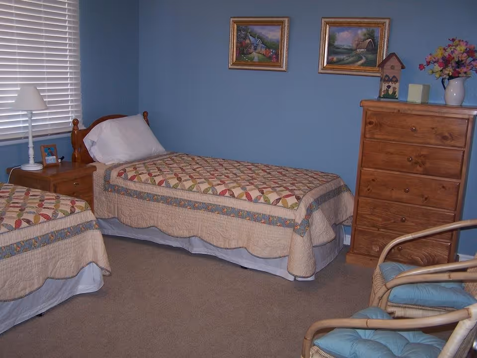 A bedroom with two twin beds covered with patterned quilts, a wooden nightstand with a white lamp and a small framed photo, a wooden chest of drawers with decorative items and a vase of flowers on top, two framed paintings on a blue wall, and two wicker chairs with blue cushions.