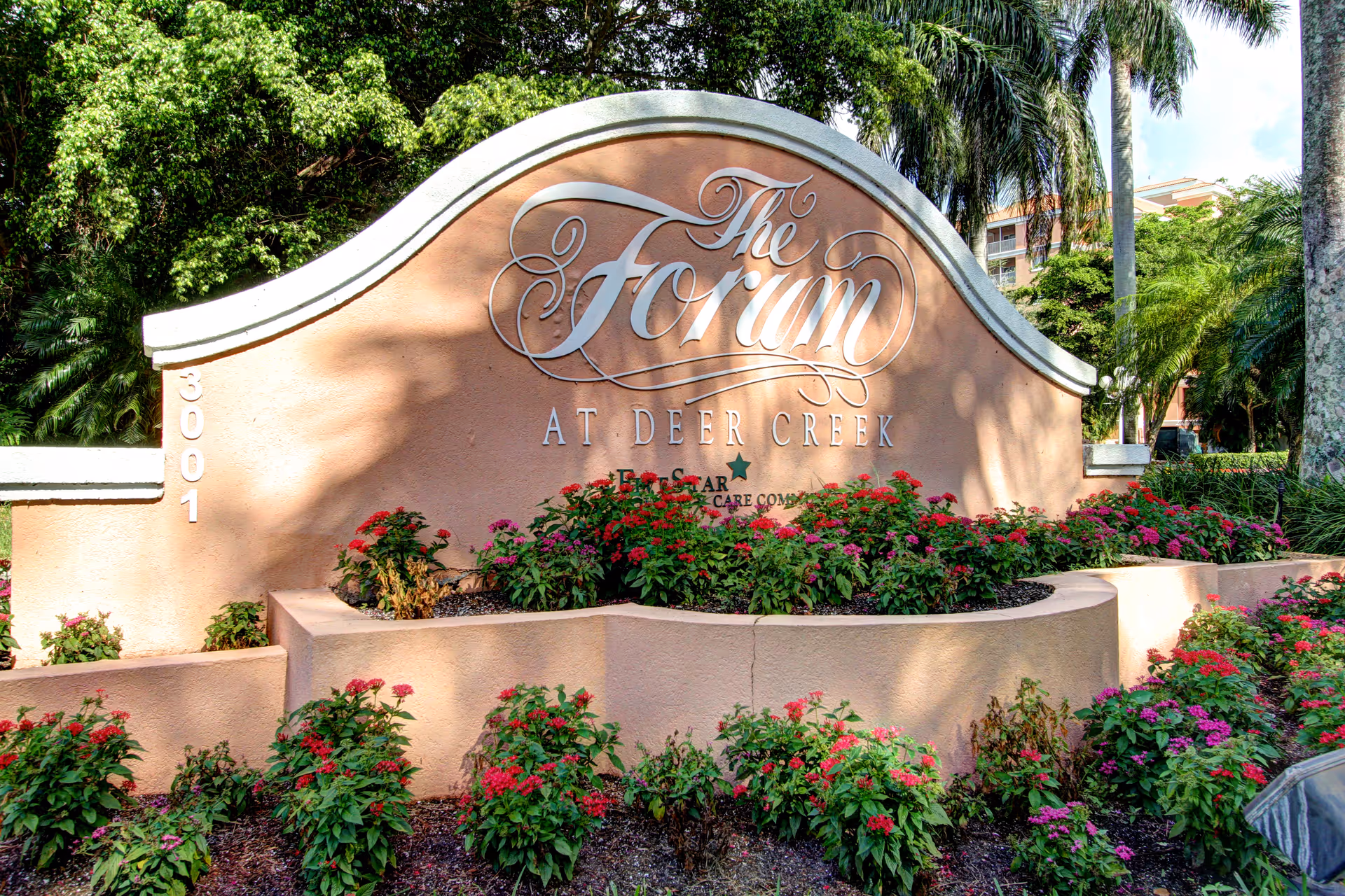 A large curved sign for The Forum at Deer Creek surrounded by colorful flowering plants and greenery, with palm trees and a building visible in the background.