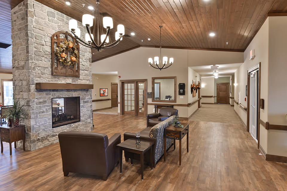 A spacious and warmly decorated common area in a senior living facility featuring a stone fireplace with a decorative wreath above it, two chandeliers hanging from a wooden ceiling, and comfortable seating including leather chairs and a patterned sofa arranged around small tables on a wood floor.