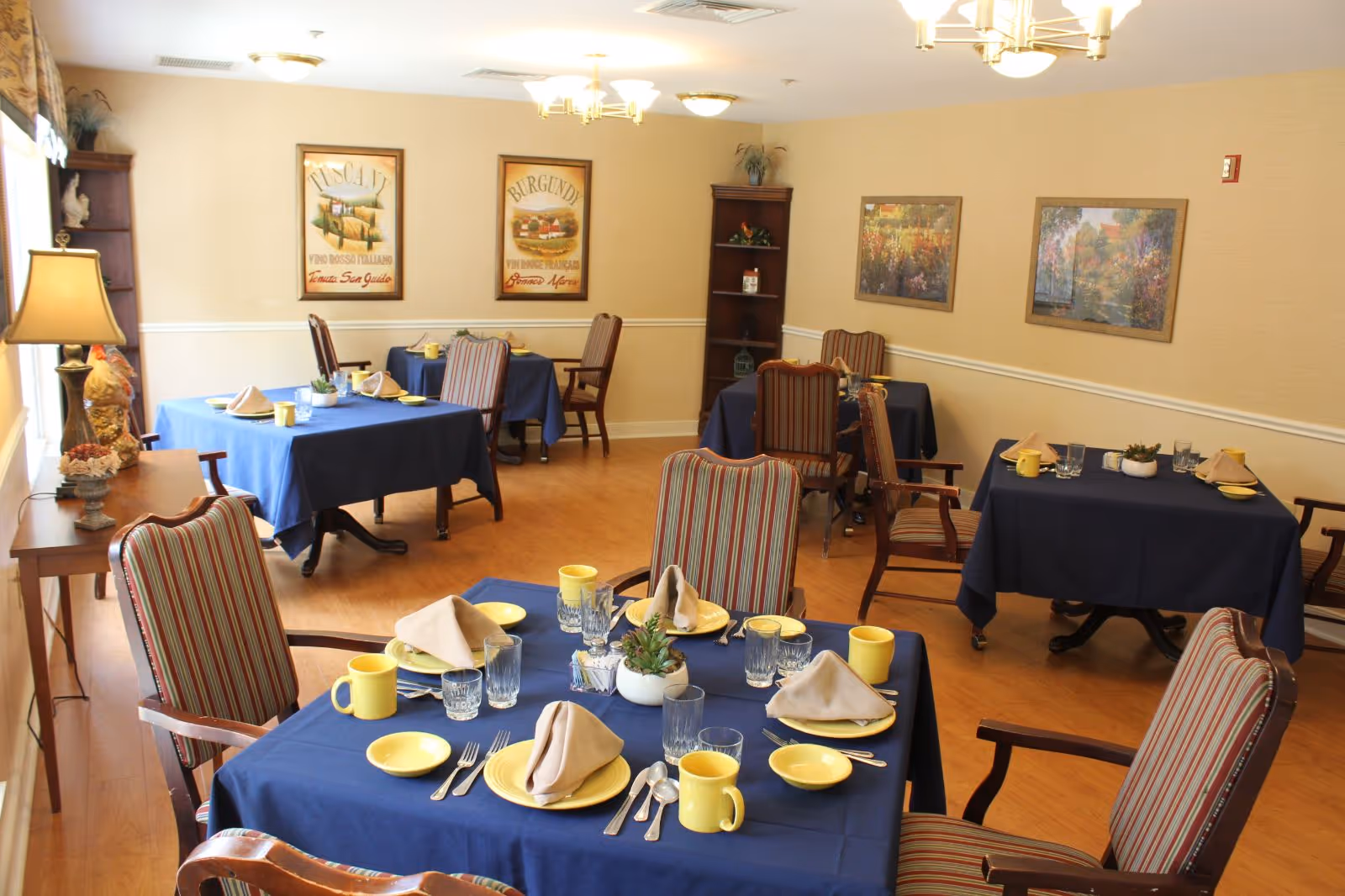A dining room with several tables covered in blue tablecloths, each set with yellow plates, cups, beige napkins, and glassware. The room has wooden floors, striped upholstered chairs, framed artwork on the walls, and a wooden shelving unit in the corner. A table lamp and decorative items are on a side table near a window with curtains.