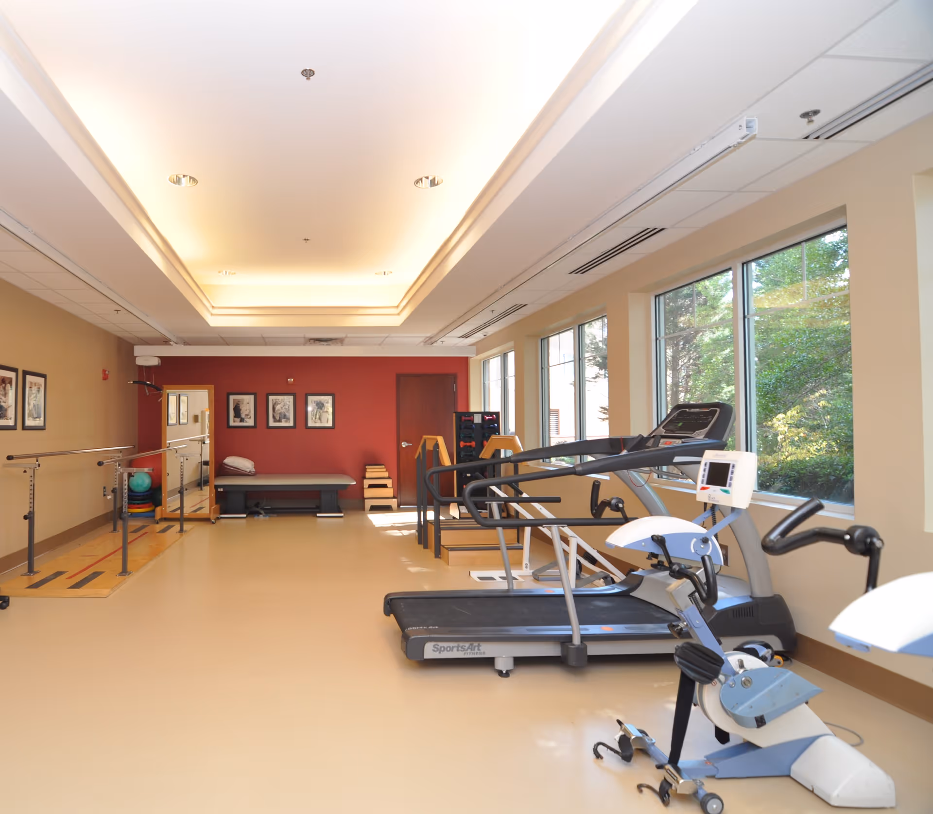 A well-lit rehabilitation or physical therapy room with exercise equipment including a treadmill, stationary bike, parallel bars for walking exercises, and a therapy bench. Large windows on the right side let in natural light and show greenery outside. The walls are painted beige and red, with framed pictures hanging on the red wall at the far end.