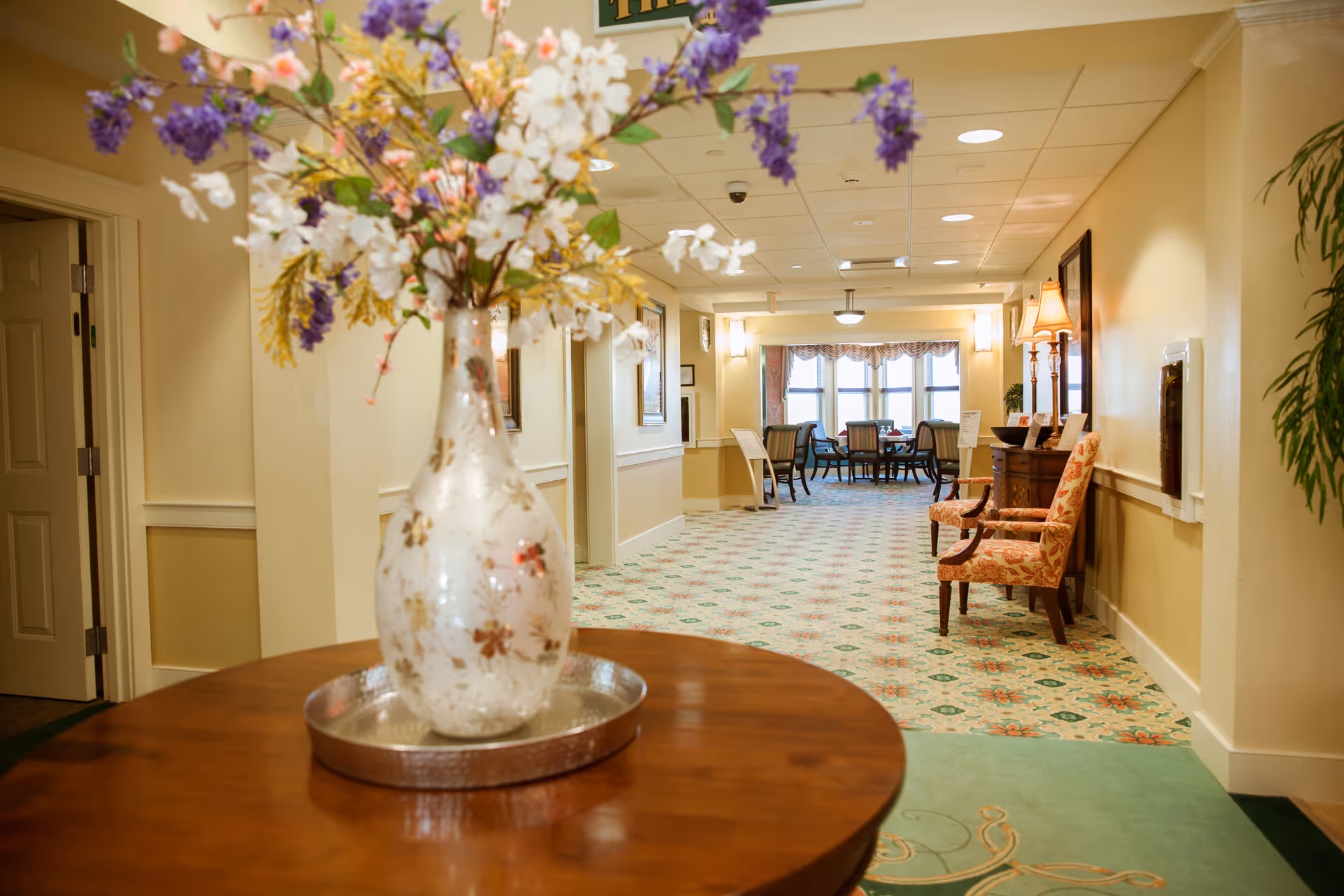 Interior hallway of Meredith Bay Colony Club featuring a round wooden table with a decorative vase of purple, white, and yellow flowers in the foreground. The hallway has patterned carpet, beige walls, framed artwork, and seating areas with upholstered chairs and lamps. At the end of the hallway, there is a window with natural light and a table with chairs.