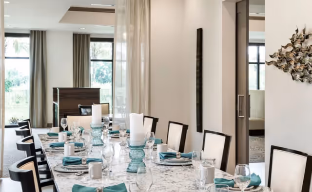 A modern dining room with a long marble table set for a meal. The table is arranged with white plates, teal napkins, glassware, and large white candles in decorative holders. The room has large windows with curtains, allowing natural light to fill the space. There is a decorative wall sculpture on the right and a glimpse of another seating area through an open doorway.