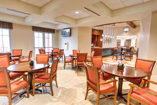 A spacious dining area with multiple round wooden tables and orange cushioned chairs. The room has large windows allowing natural light to brighten the space. There are small flower arrangements on each table and a television mounted on the wall. The area extends into another dining or lounge space with additional seating.