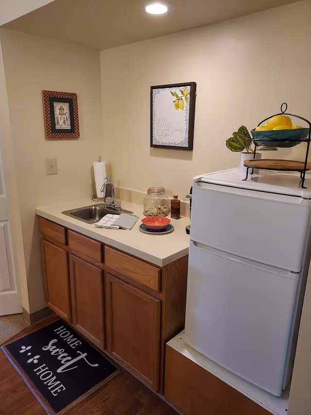 Small kitchenette area with a white mini refrigerator topped with a two-tiered fruit basket containing lemons and a plant. Next to the refrigerator is a countertop with a sink, paper towel holder, a jar of cookies, a small red bowl on a plate, and a bottle of seasoning. Two framed pictures hang on the beige wall above the counter. A black floor mat with the words 'Home Sweet Home' is placed on the wooden floor in front of the cabinets.
