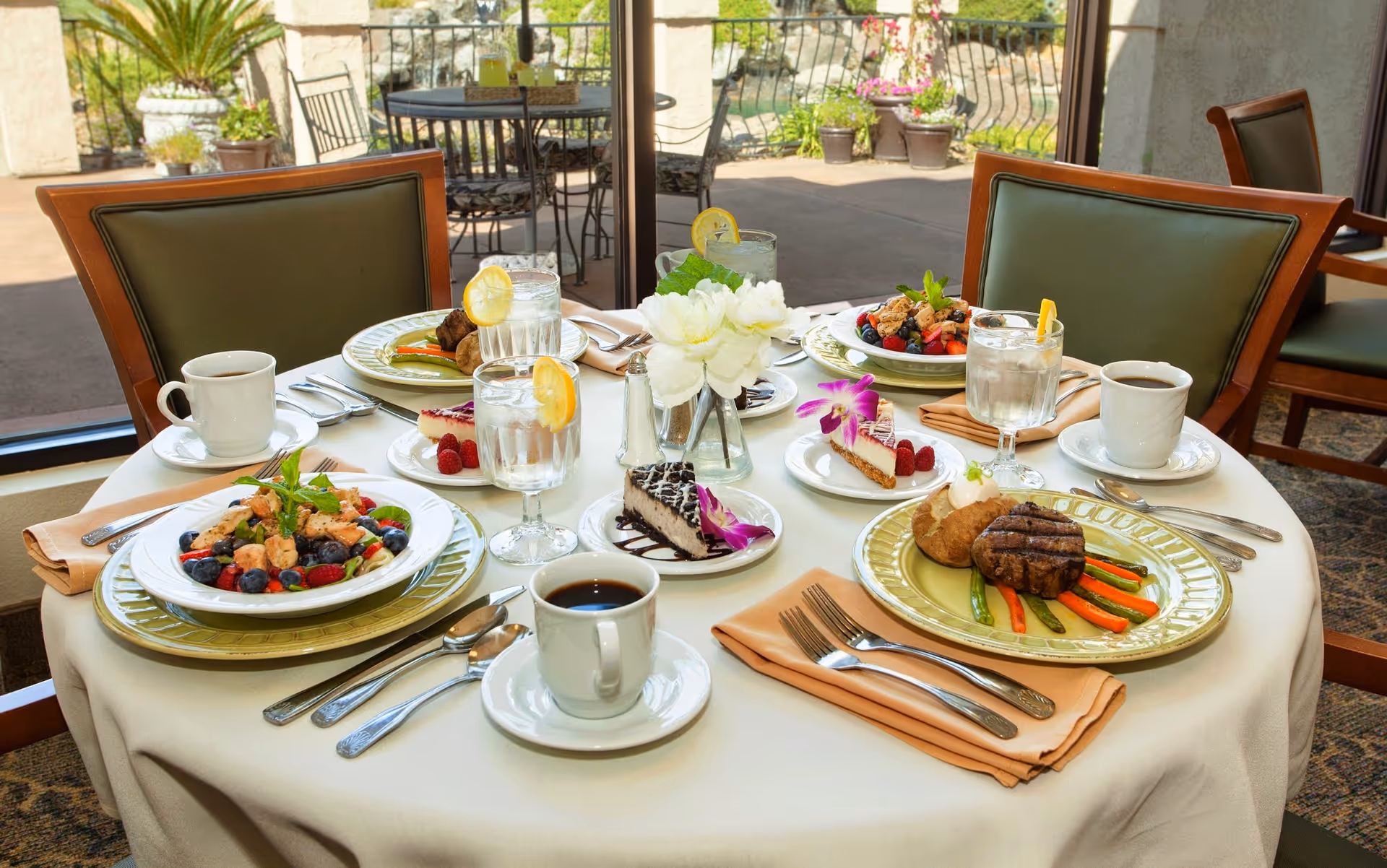 Table in a dining room set for four with plated entrees, desserts, coffee cups, and glasses by a window overlooking a patio.