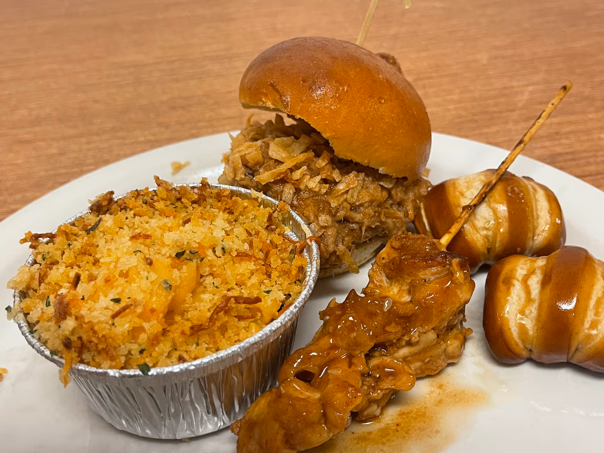 A white plate with a small sandwich filled with crispy fried onions, a foil cup of baked macaroni and cheese topped with breadcrumbs, a skewer of glazed grilled chicken, and two soft pretzels drizzled with sauce, all placed on a wooden table.