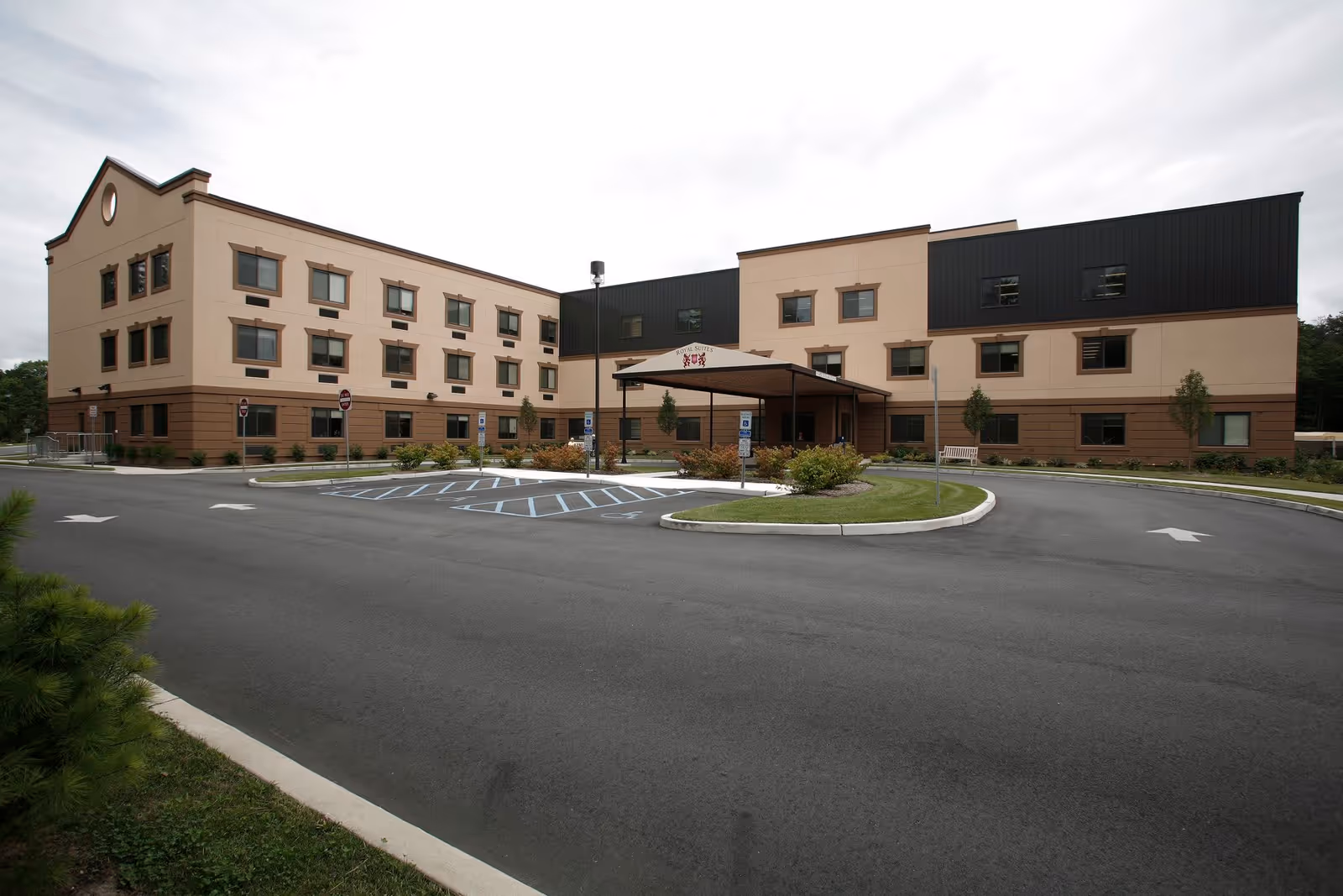 Exterior view of a three-story beige and brown building with multiple windows, a covered entrance with a canopy labeled 'Royal Suites', surrounded by a paved driveway and parking spaces including handicapped spots, under a cloudy sky.