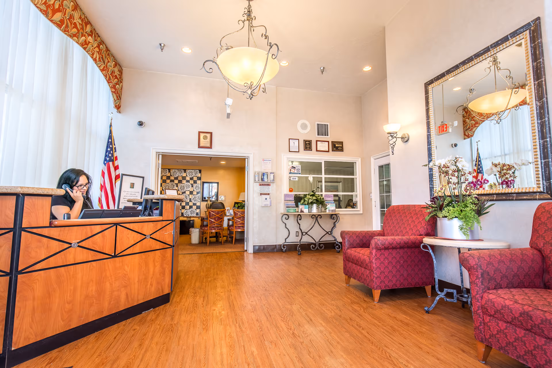 Reception area of a healthcare facility with a woman sitting behind a wooden front desk talking on the phone. The room has wooden flooring, two red upholstered armchairs with a small round table and a flower arrangement between them, a large mirror on the wall, and an American flag near a window with sheer white curtains. There is a doorway leading to another room with a dining area visible in the background.