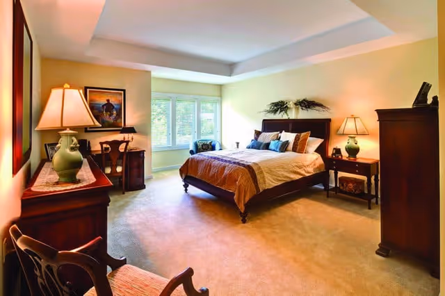 Bright, spacious bedroom with a large bed, nightstands and lamps, dresser, and a seating area by a window.