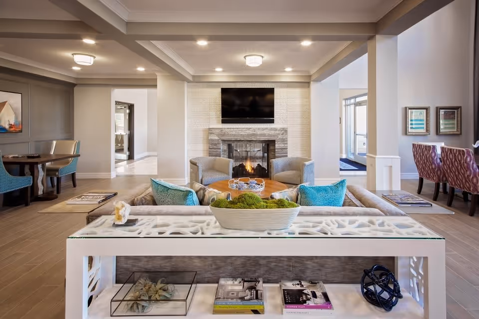 A modern and cozy living room area in a senior living facility featuring a fireplace with a mounted TV above it, two armchairs facing the fireplace, a sofa with turquoise pillows, and a white console table with decorative items and books. The room has neutral tones with wooden flooring and recessed lighting, and there are additional seating areas with tables and chairs visible in the background.