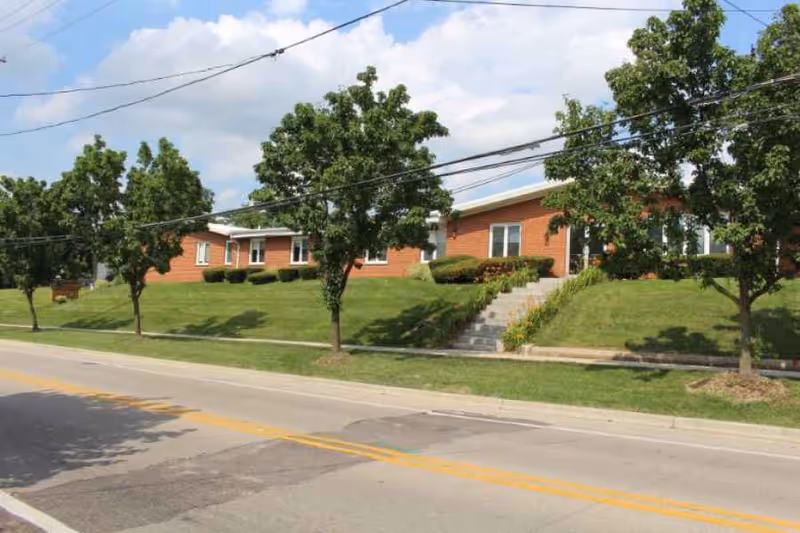 Exterior view of a single-story brick building with a sloped grassy lawn and several trees in front. A concrete staircase leads up to the entrance of the building from the sidewalk along a paved road.