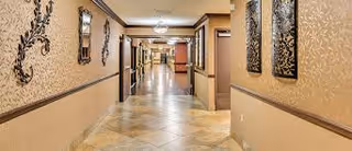 A long, well-lit hallway in an assisted living facility with beige walls decorated with ornate wall hangings and mirrors. The floor is tiled with a light-colored pattern, and there are multiple doorways along the corridor.