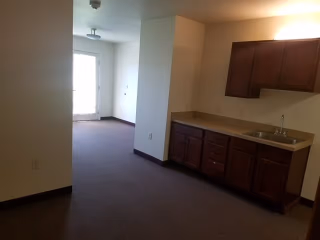 Empty interior room with a kitchenette (sink and cabinets) on the right and a door with window at the far end.