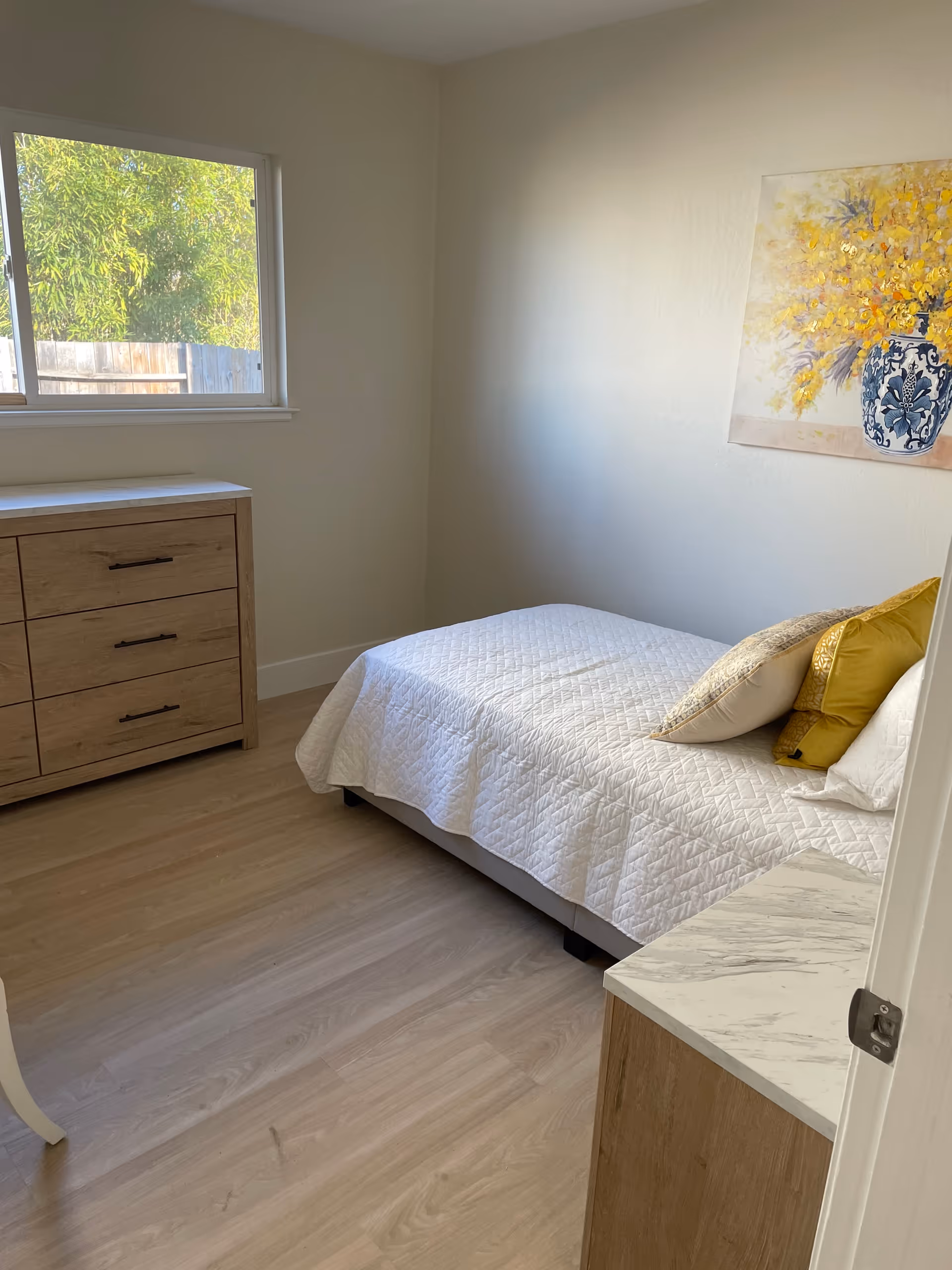 A small bedroom with a single bed covered with a white quilt and several pillows in white and yellow. There is a wooden dresser with three drawers against the wall under a window showing greenery outside. A painting of yellow flowers in a blue and white vase hangs on the wall above the bed. The floor is light wood, and a small marble-topped nightstand is partially visible in the foreground.