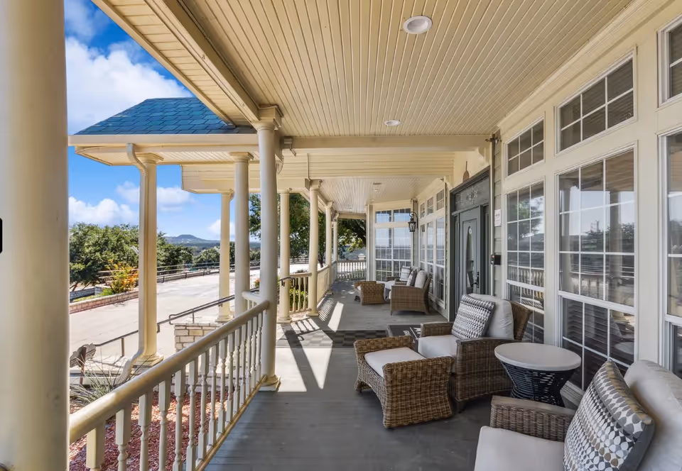 Covered porch with wicker seating and small tables along a columned railing overlooking a landscaped drive and distant hills.