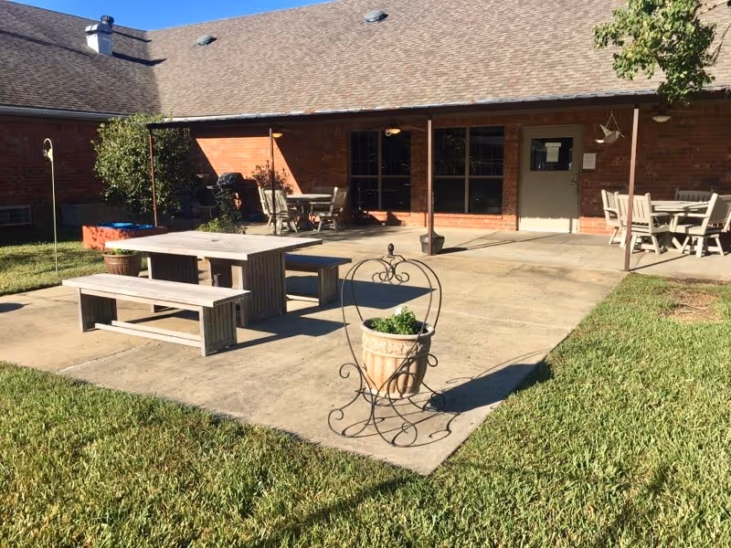 Outdoor patio area at Copiah Living Center featuring wooden picnic tables and benches on a concrete slab, surrounded by green grass and a brick building with a covered porch in the background. A decorative metal plant holder with a potted plant is in the foreground.