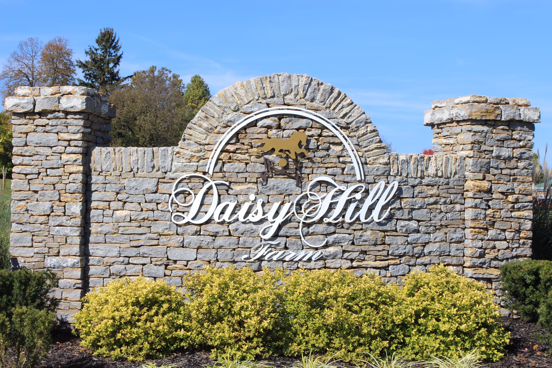 Stone entrance sign for Daisy Hill Farm with a decorative arch and a horse silhouette above the name, surrounded by green shrubs and trees under a clear blue sky.