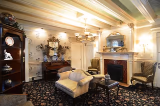A cozy living room with vintage decor featuring a patterned carpet, a beige loveseat, two upholstered armchairs, a wooden coffee table, a fireplace with a mirror above it, a piano against the wall, and a chandelier hanging from the ceiling.