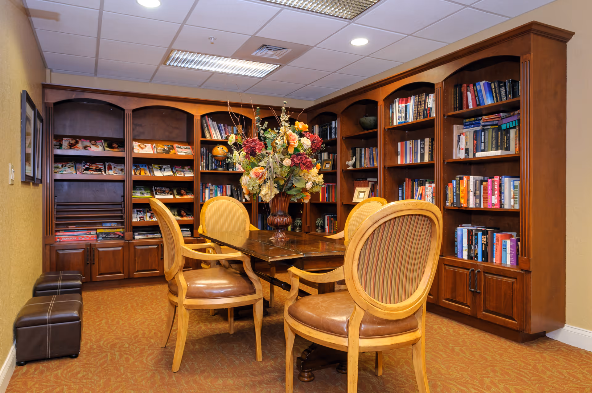 Wood-paneled library/reading room with bookshelves, a wooden table surrounded by upholstered chairs and a large floral centerpiece.