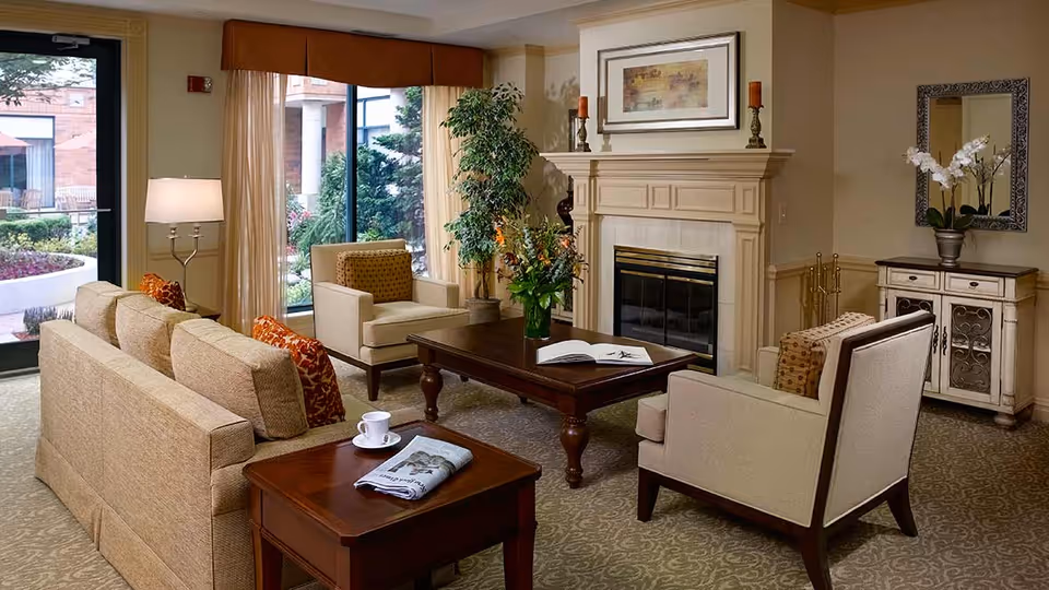 A cozy living room area in a senior living facility with beige sofas and armchairs arranged around a wooden coffee table. The room features a fireplace with decorative candlesticks and a framed picture above it. There is a side table with a cup and newspaper, a floor lamp, a large window with curtains showing an outdoor garden, a potted plant, and a cabinet with a mirror and orchid flowers.