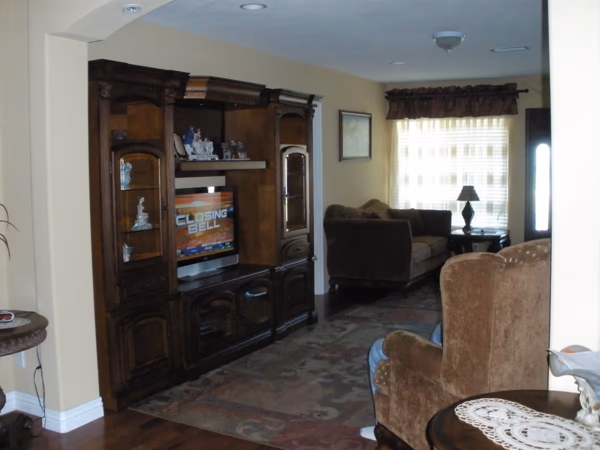 A living room with a large wooden entertainment center holding a television displaying the text 'Closing Bell.' The room has beige walls, a patterned area rug, a brown upholstered armchair in the foreground, and a brown sofa near a window with sheer curtains and a valance. A small table with a lamp is next to the sofa.