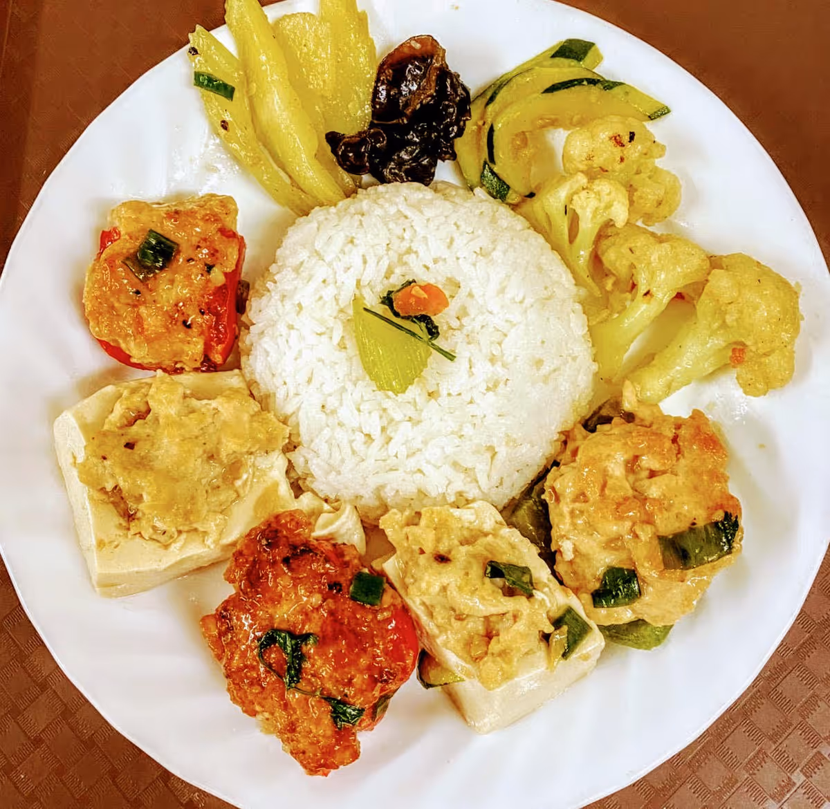 A white plate with a serving of white rice in the center, surrounded by various cooked vegetables including cauliflower, zucchini, and pieces of tofu topped with a savory sauce.