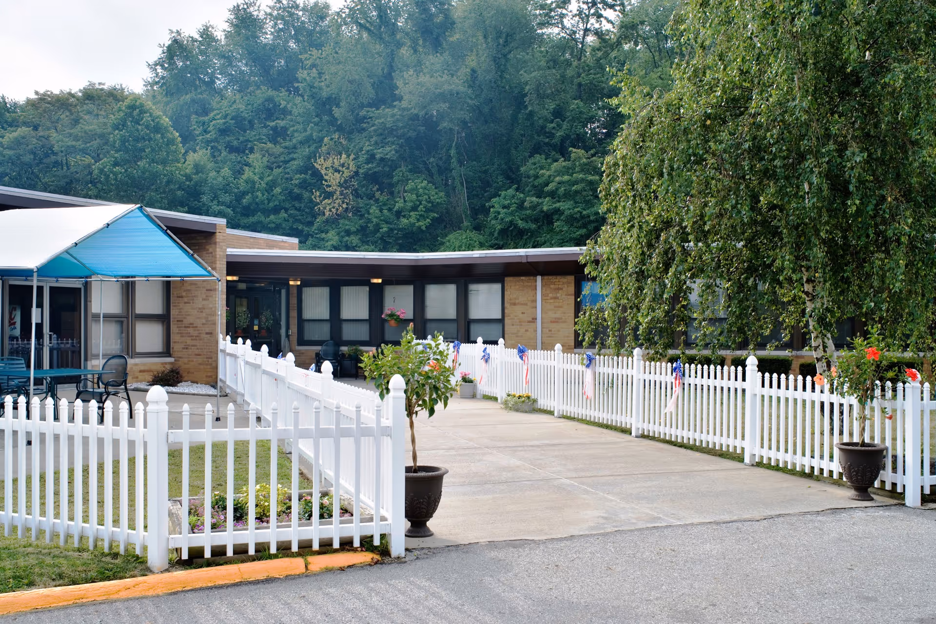 Outdoor view of Laurel Ridge Center showing a paved walkway lined with white picket fences decorated with small American flags. There are potted plants on either side of the walkway and a blue canopy covering a seating area with tables and chairs. The building is single-story with large windows and surrounded by trees and greenery.