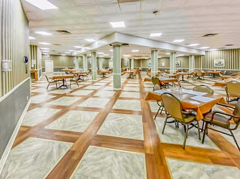A spacious dining room with multiple tables covered with orange tablecloths and chairs arranged around them. The room has a patterned floor with wood and tile sections, striped wallpaper, and several columns supporting the ceiling. The lighting is bright with recessed ceiling lights, and framed pictures hang on the walls.