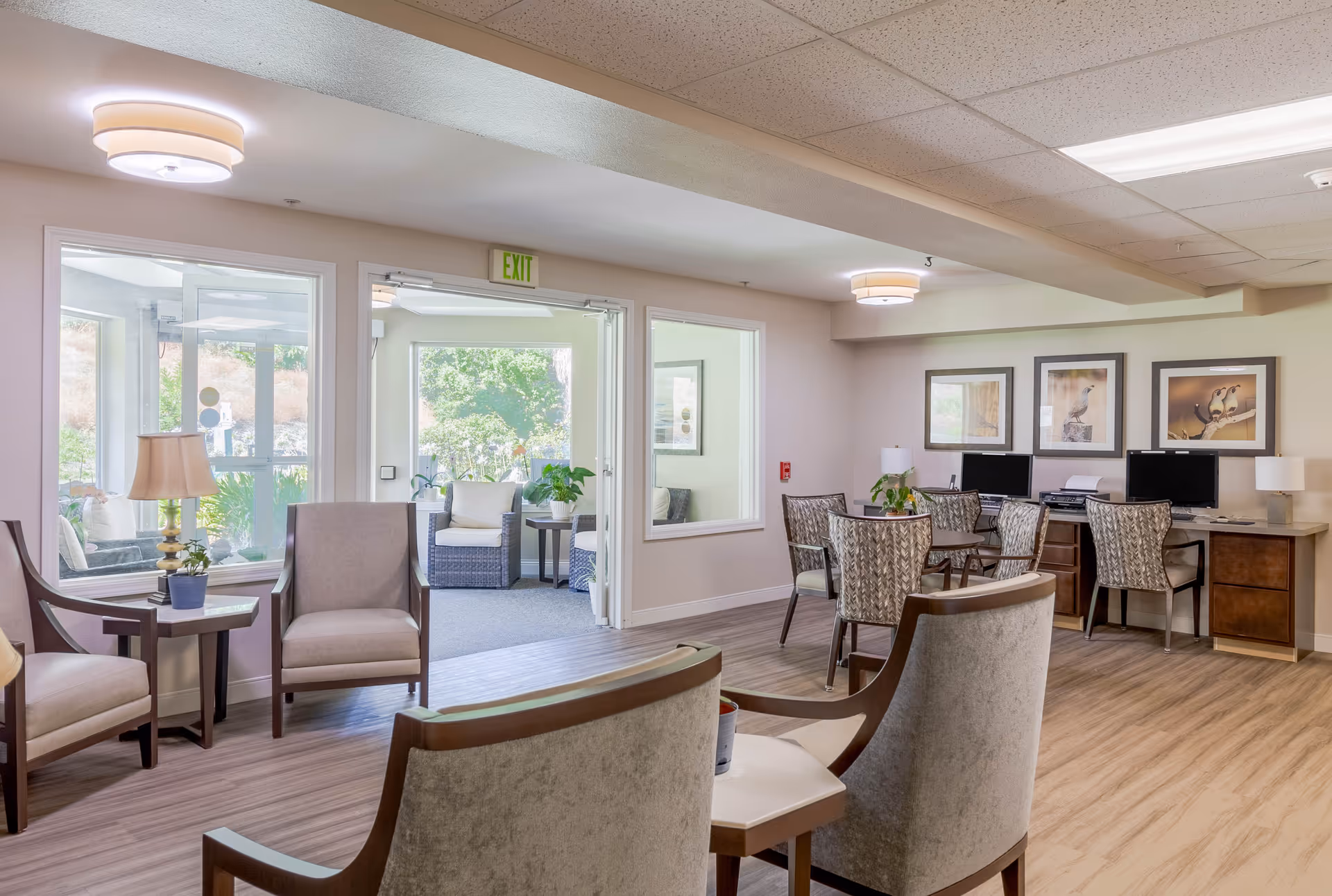 Bright communal lounge in a retirement community with armchairs, tables, computers, and a seating area by large windows overlooking greenery.