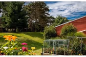 Outdoor garden area with colorful flowers in the foreground, a raised garden bed with plants, green grass, trees, and a red building under a partly cloudy sky.