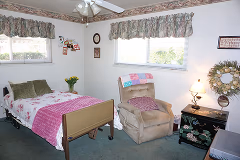 A bright senior bedroom with an adjustable bed, a recliner, bedside table and two windows with floral valances.