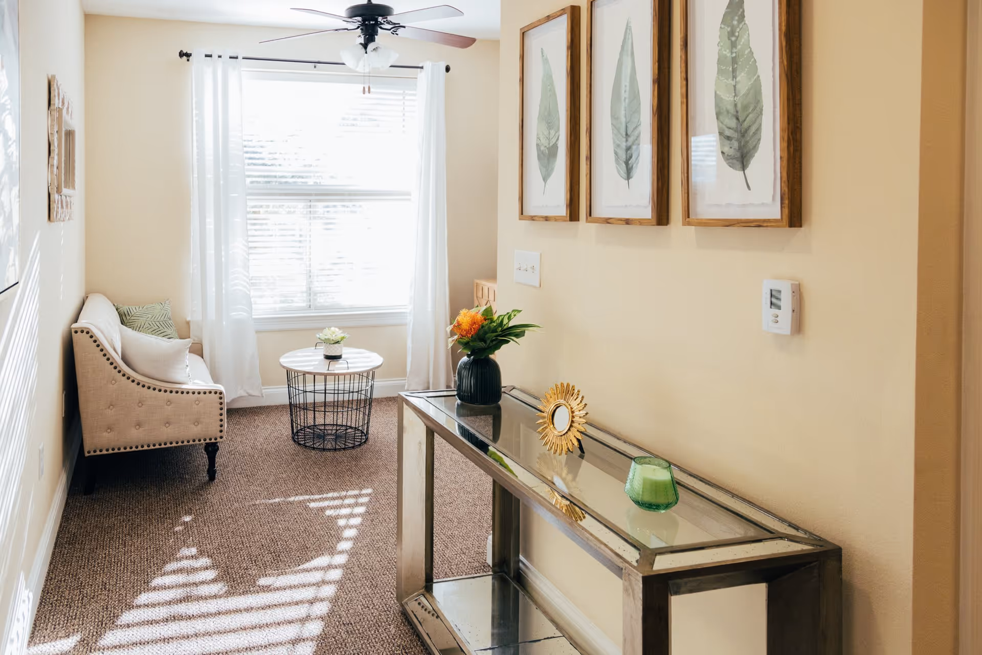 A bright and cozy living room area with a beige upholstered loveseat adorned with pillows, a round wire-frame coffee table with a small plant on top, and a glass console table holding a vase with flowers, a decorative sunburst mirror, and a green candle. Three framed leaf prints hang on the wall above the console table, and a ceiling fan with lights is mounted on the ceiling. Sunlight streams through a window with white curtains, casting shadows on the carpeted floor.