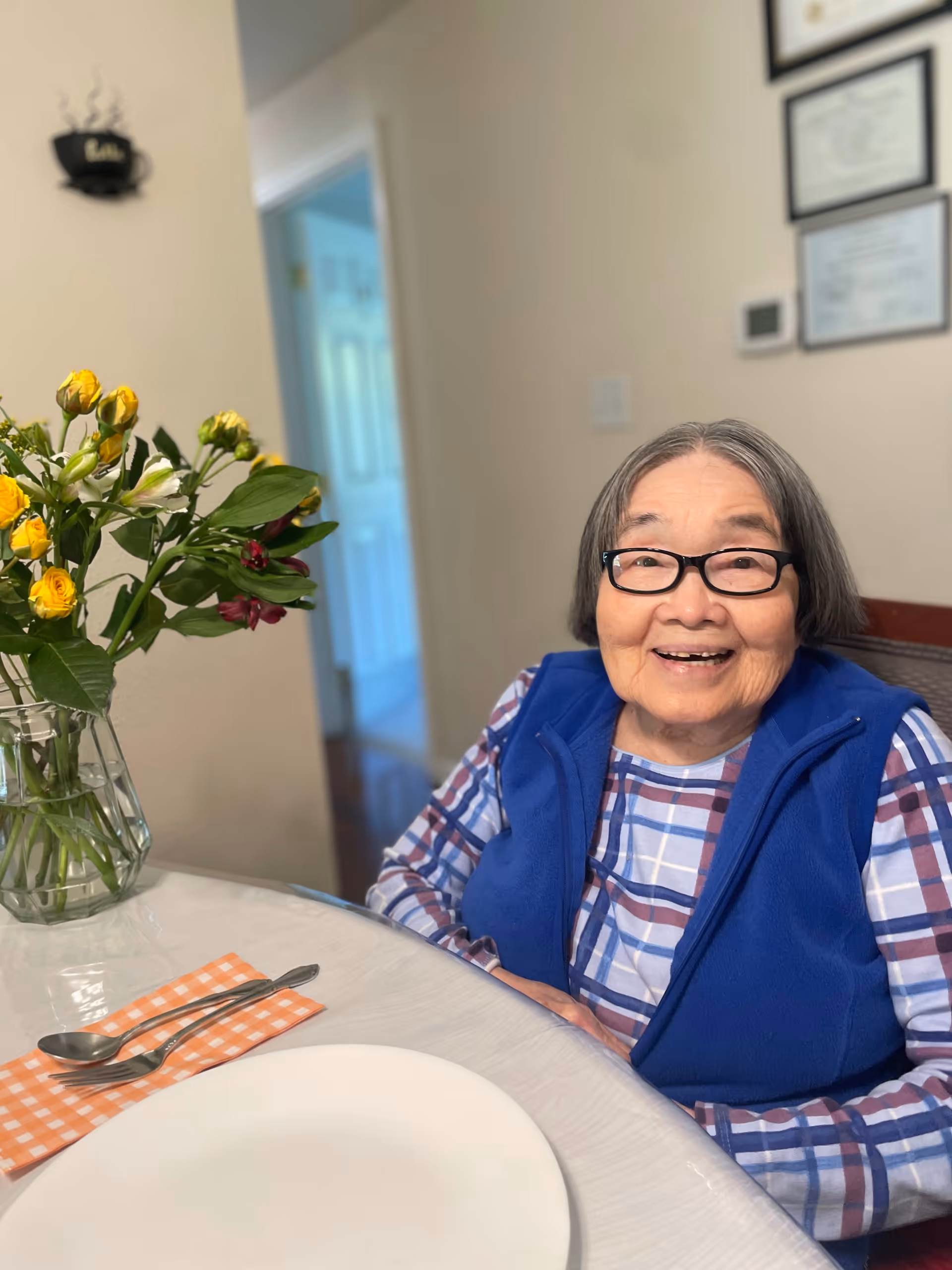 An elderly woman with glasses and short gray hair is sitting at a dining table, smiling. She is wearing a blue vest over a plaid shirt. On the table in front of her is a white plate, a fork and spoon on an orange checkered napkin, and a vase with yellow and white flowers. The background shows a hallway and some framed certificates on the wall.