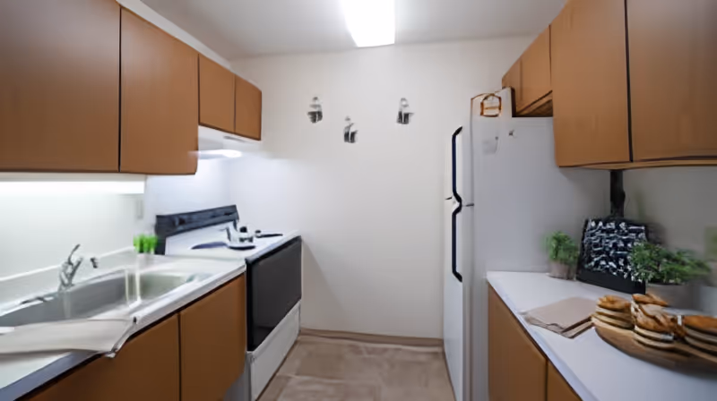 A small kitchen with brown cabinets, a white refrigerator, a stove, a double sink, and white countertops. There are some green plants and a plate with stacked sandwiches on the counter. The walls are white with three small decorative wall hangings.