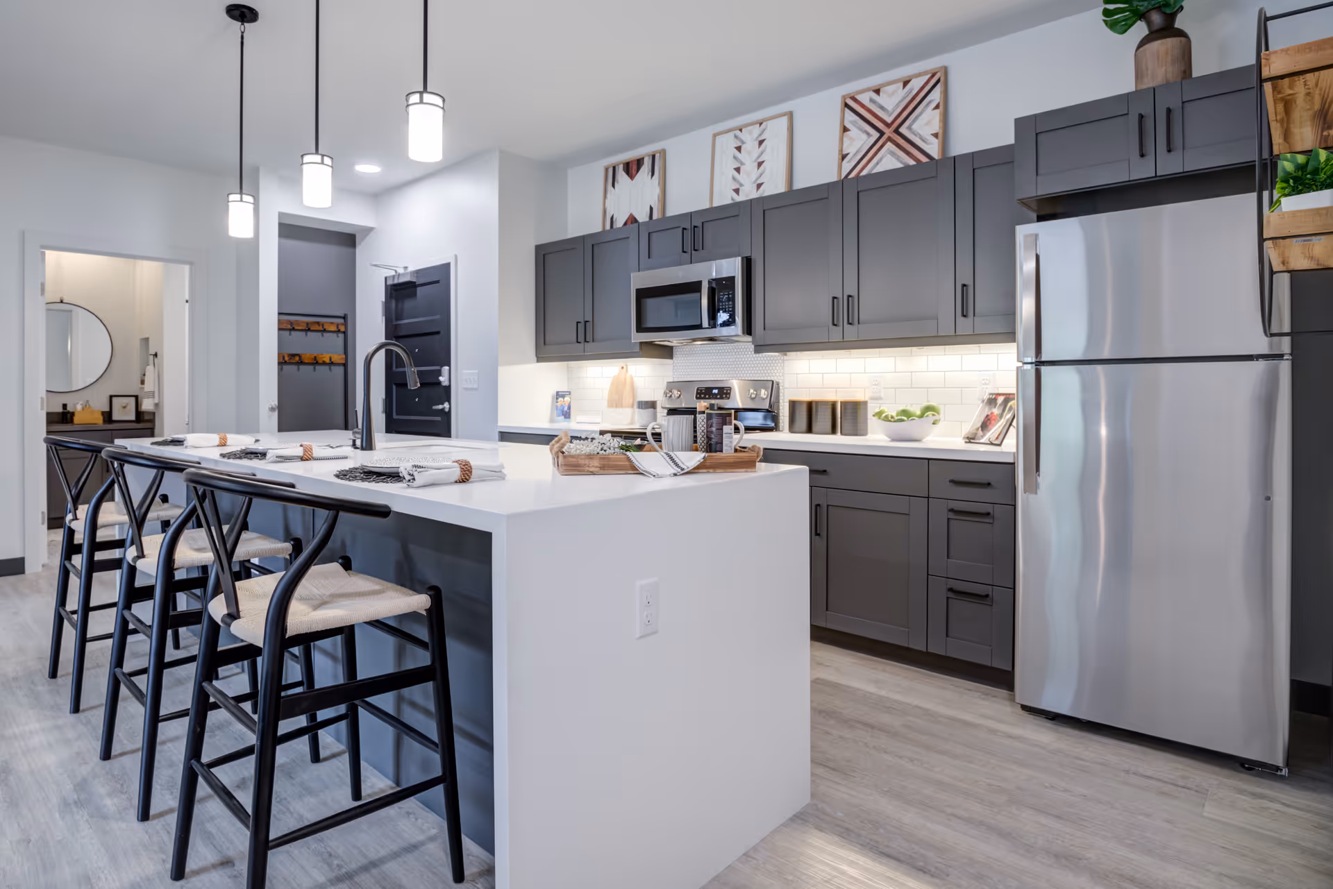Modern kitchen with a large white island featuring four black chairs with woven seats. The kitchen has gray cabinets, a stainless steel refrigerator, microwave, and stove. Three pendant lights hang above the island. Decorative framed artwork is displayed above the cabinets, and the floor is light wood.