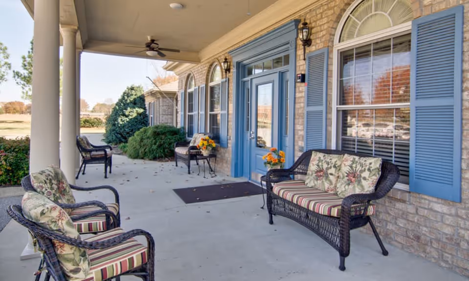 Covered outdoor patio area at Greenbrier Meadows with wicker chairs and a loveseat featuring floral and striped cushions. The building has brick walls, blue shutters, and a blue door with glass panels. There are potted flowers on small tables and bushes in the background.