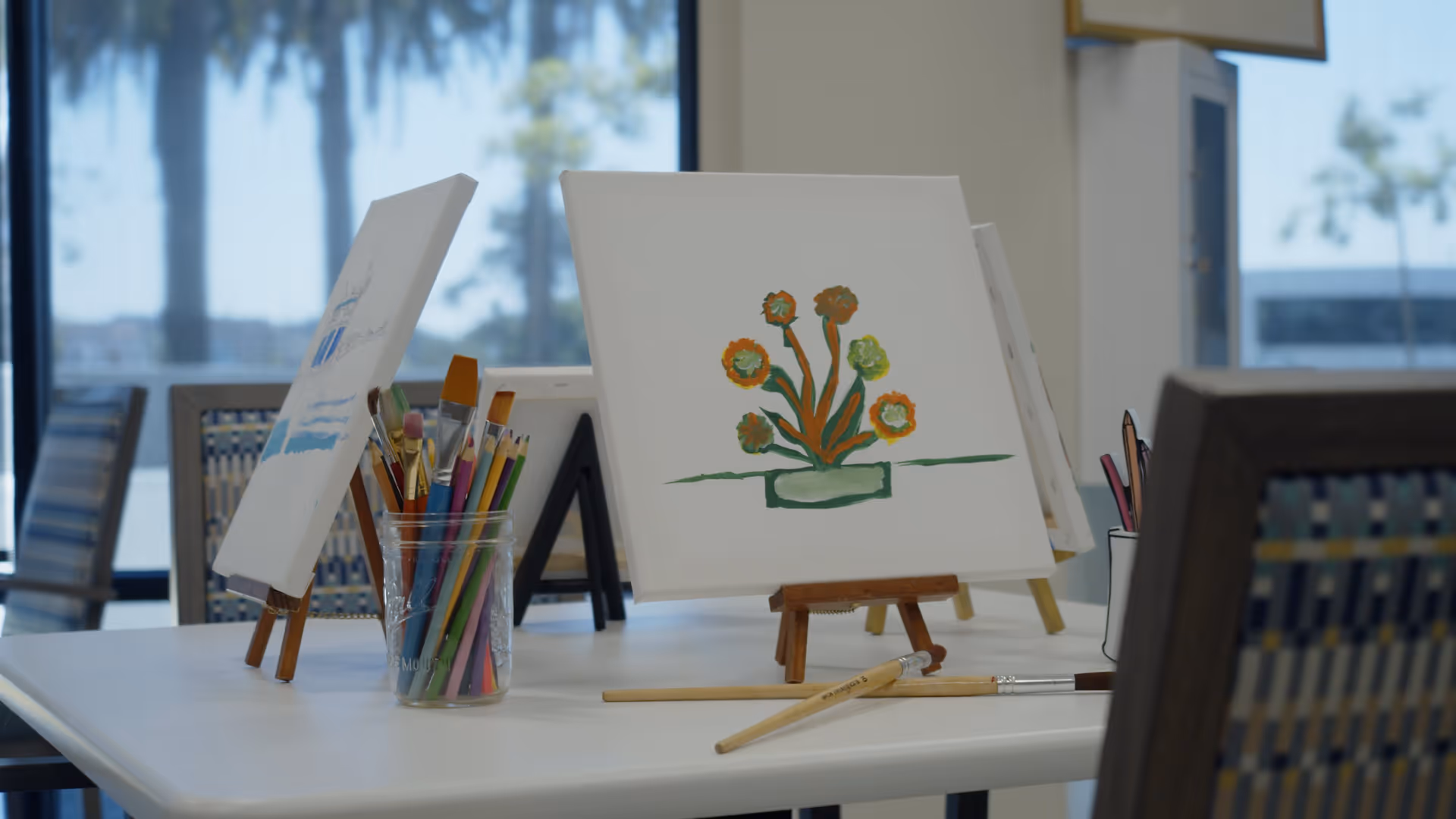 A table with small easels holding canvases displaying paintings, including a floral painting. There are paintbrushes and colored pencils in clear containers on the table. The background shows large windows with a view of palm trees outside.