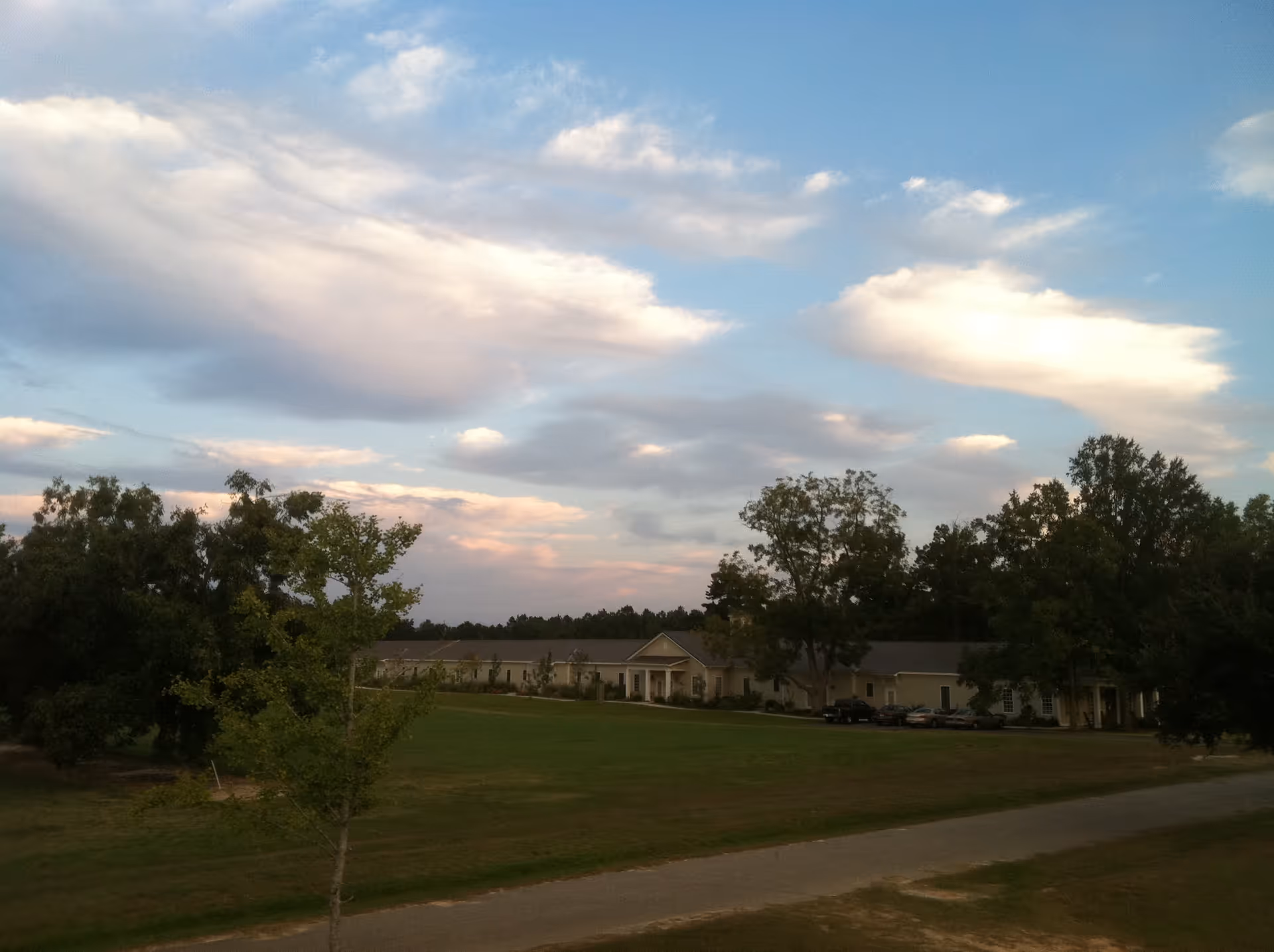 A wide view of Fellowship Brookside Assisted Living Community building with a large green lawn in front, surrounded by trees under a partly cloudy sky during late afternoon or early evening.
