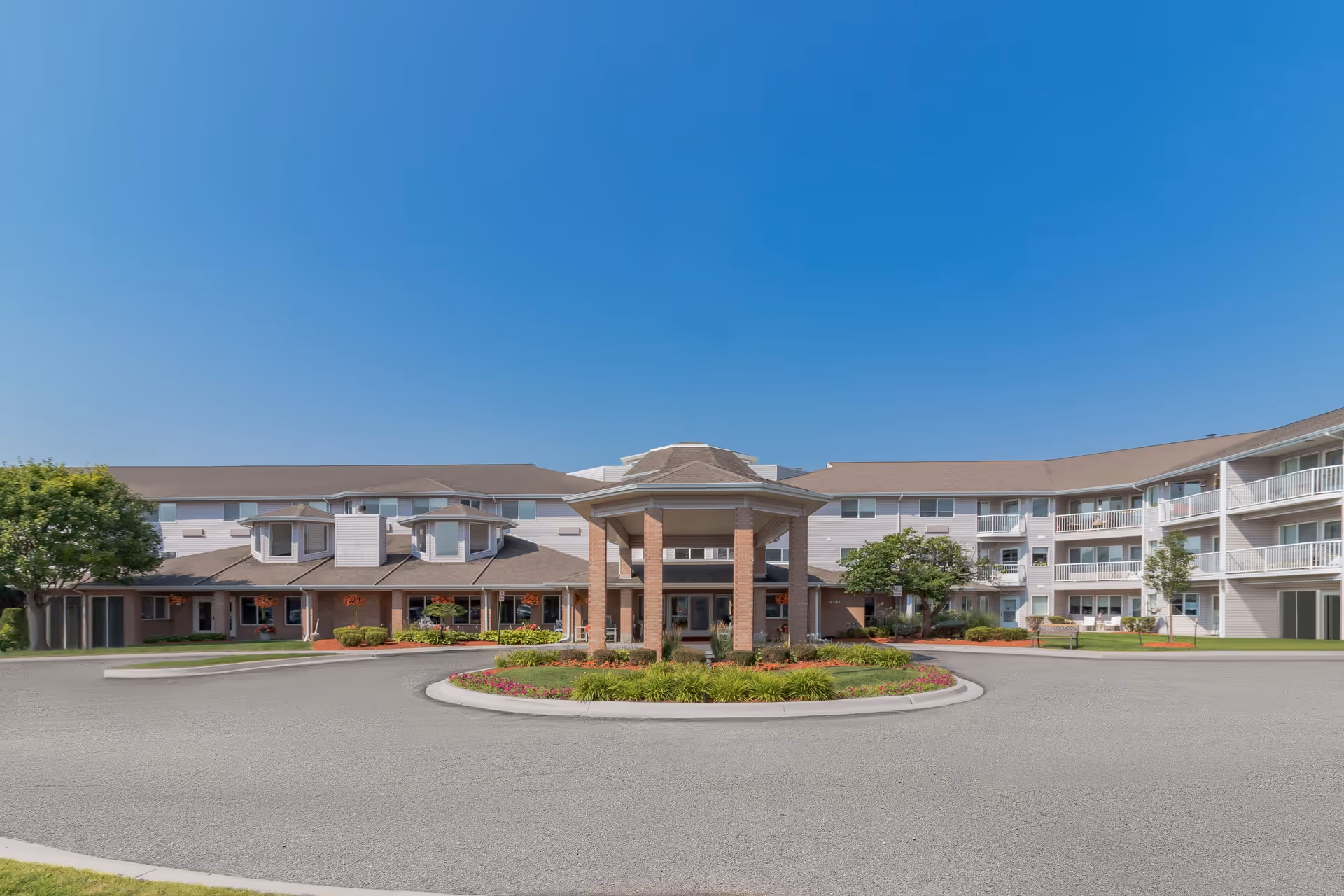 Front exterior view of Independence Village of Saginaw, a multi-story senior living facility with a circular driveway and a covered entrance. The building has balconies, windows, and landscaped greenery with trees and flowers under a clear blue sky.