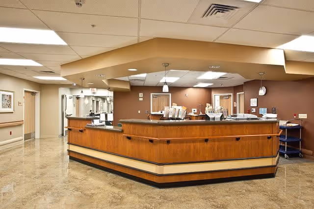 A central wooden nurses' station and reception desk in a well-lit nursing facility hallway.