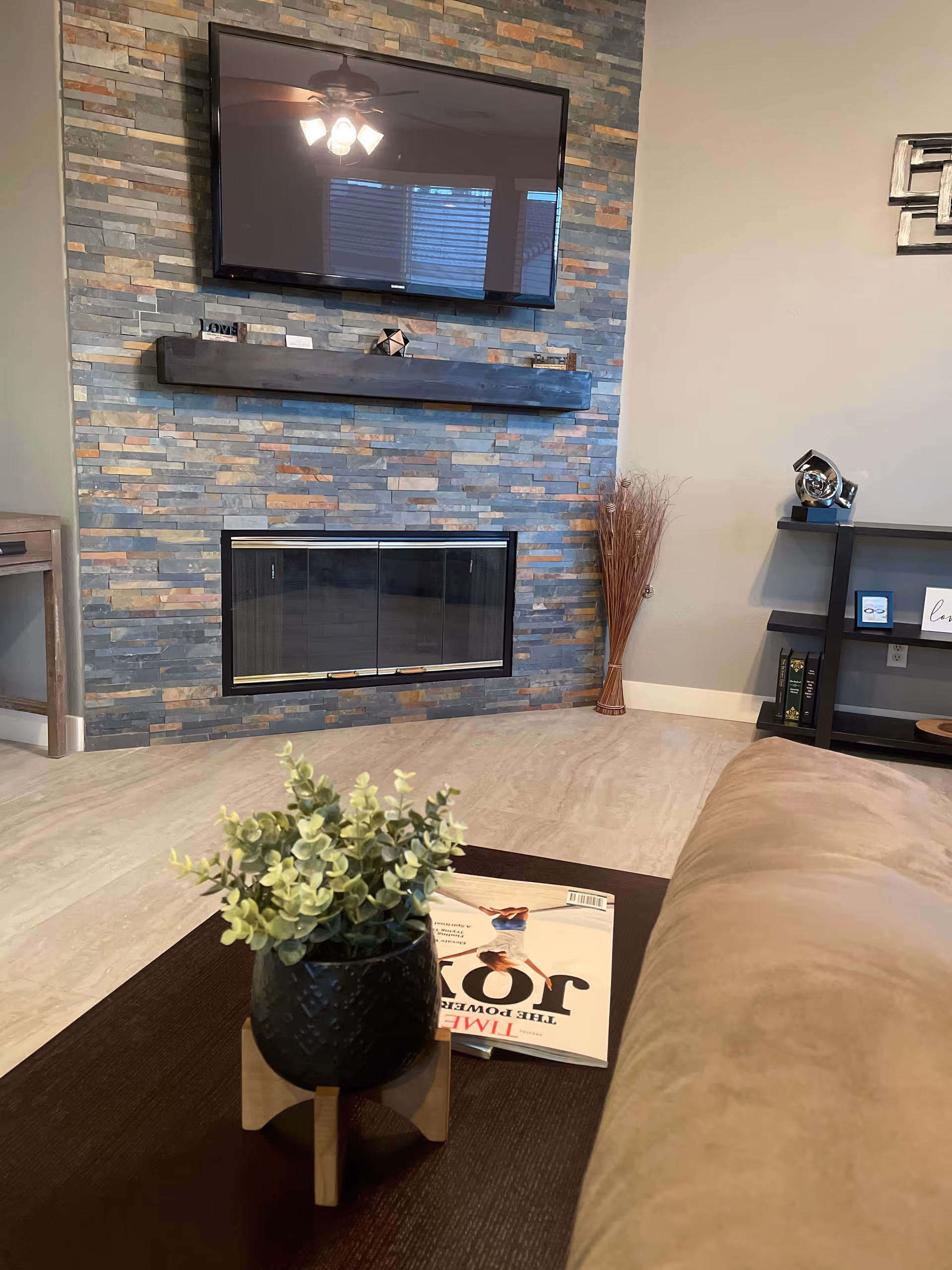 A cozy living room with a stone accent wall featuring a mounted flat-screen TV above a modern fireplace. In the foreground, there is a dark wooden coffee table with a small potted plant and a magazine on it. To the right, a beige couch is partially visible, and a black shelving unit with decorative items is against the wall.