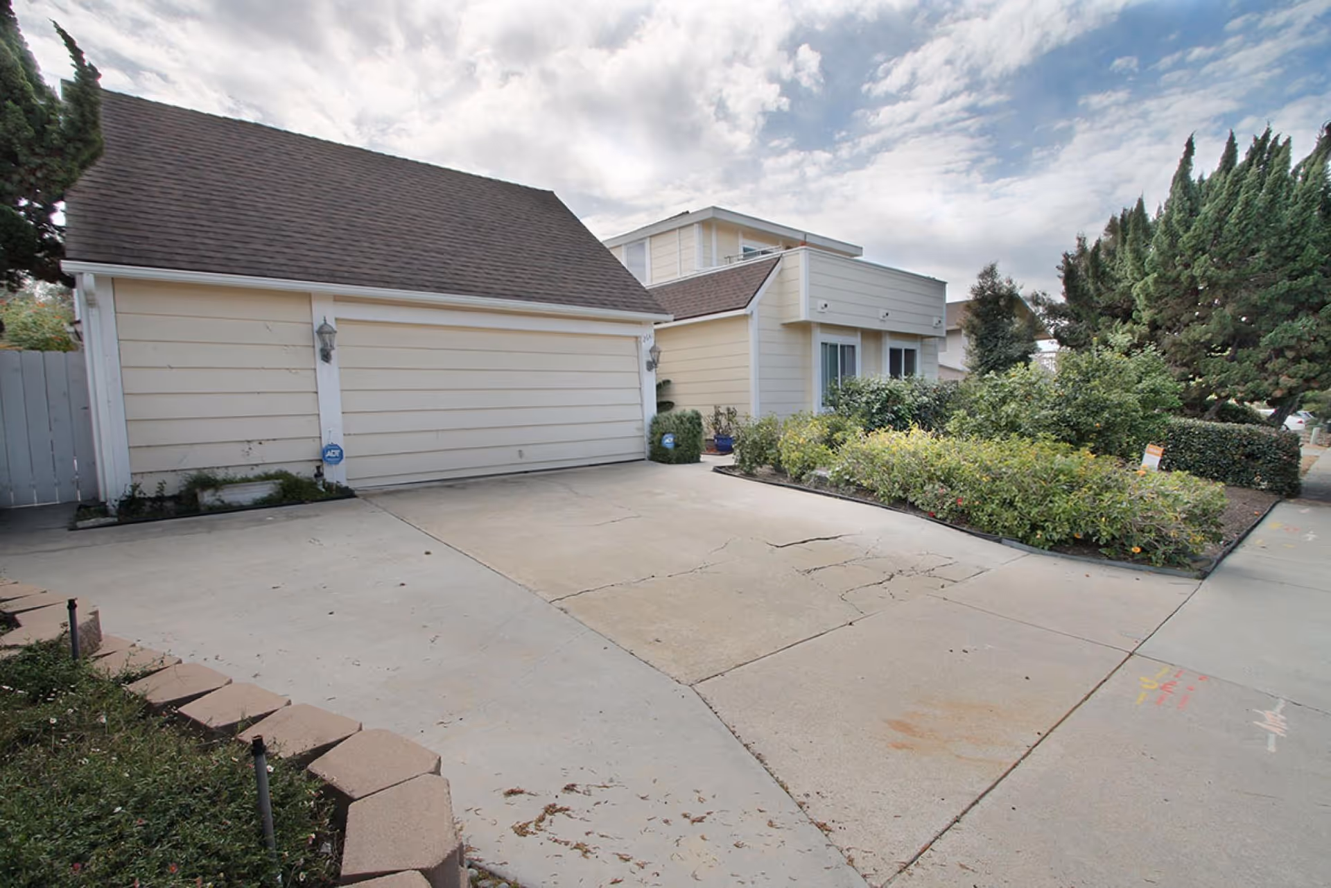 Driveway and double garage of a light-yellow, two-story house with landscaped front beds under a cloudy sky.